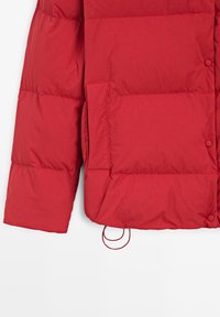 Manche et partie basse du torse d'une veste matelassée rouge avec boutons-pression, poche latérale et cordon de serrage réglable visibles sur fond blanc.