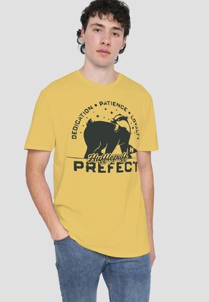 Junger Mann trägt ein gelbes Hufflepuff-Prefekt-T-Shirt mit einem schwarzen Dachs und den Worten Hingabe, Geduld, Loyalität.
