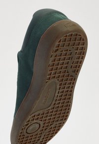 Zöld velúr sportcipő sötét gumi talppal, texturált mintázattal és kör alakú díszít�éssel. A Lacoste logó a talpba van nyomva.