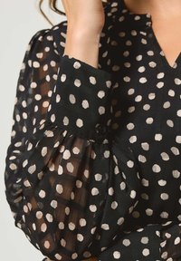 Blusa nera in tessuto trasparente, con grandi pois beige. Abbottonatura frontale con scollo a V e maniche a sbuffo arricciate che terminano con un polsino abbottonato.