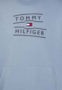 Felpa leggera blu con cappuccio, con il testo nero "Tommy Hilfiger" e il logo rettangolare rosso, bianco e blu del marchio sopra la tasca anteriore.