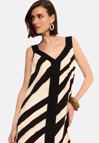 Długa, bezrękawna sukienka z czarno-kremowym wzorem w ukośne paski. Dekolt w kształcie litery V, dopełniony grubą bransoletką.