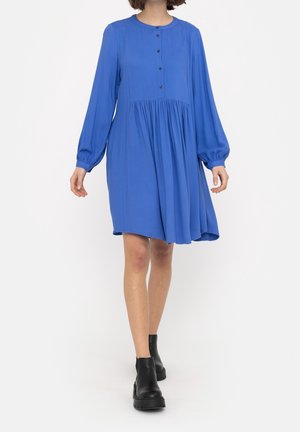 Kobieta w luźnej, niebieskiej sukience z długimi rękawami i czarnych botkach, idąca na tle jednolitego białego tła.