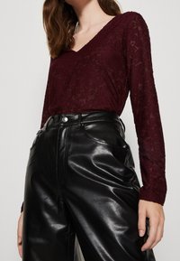 Burgunderfarbene Spitzenbluse mit langen Ärmeln und V-Ausschnitt, kombiniert mit hochgeschnittenen schwarzen Lederhosen mit zwei Vordertaschen und glänzender Textur.
