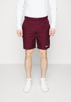 Hombre con pantalones cortos deportivos Nike granate, camisa blanca de manga larga y zapatillas Nike blancas, de pie frente a un fondo liso.