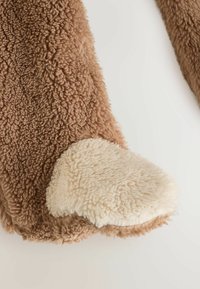 Braunes Kunstfellmaterial mit einem weichen, elfenbeinfarbenen pfotenartigen Design. Strukturiert, flauschige Oberfläche. Abgerundete Form mit kontrastierenden Farbdetails.