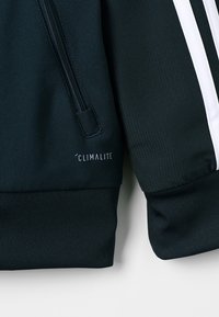Mørkeblå atletisk jakke med lynlåslomme, ribbede manchetter og hvide striber, med 'CLIMALITE' branding synlig på stoffet.