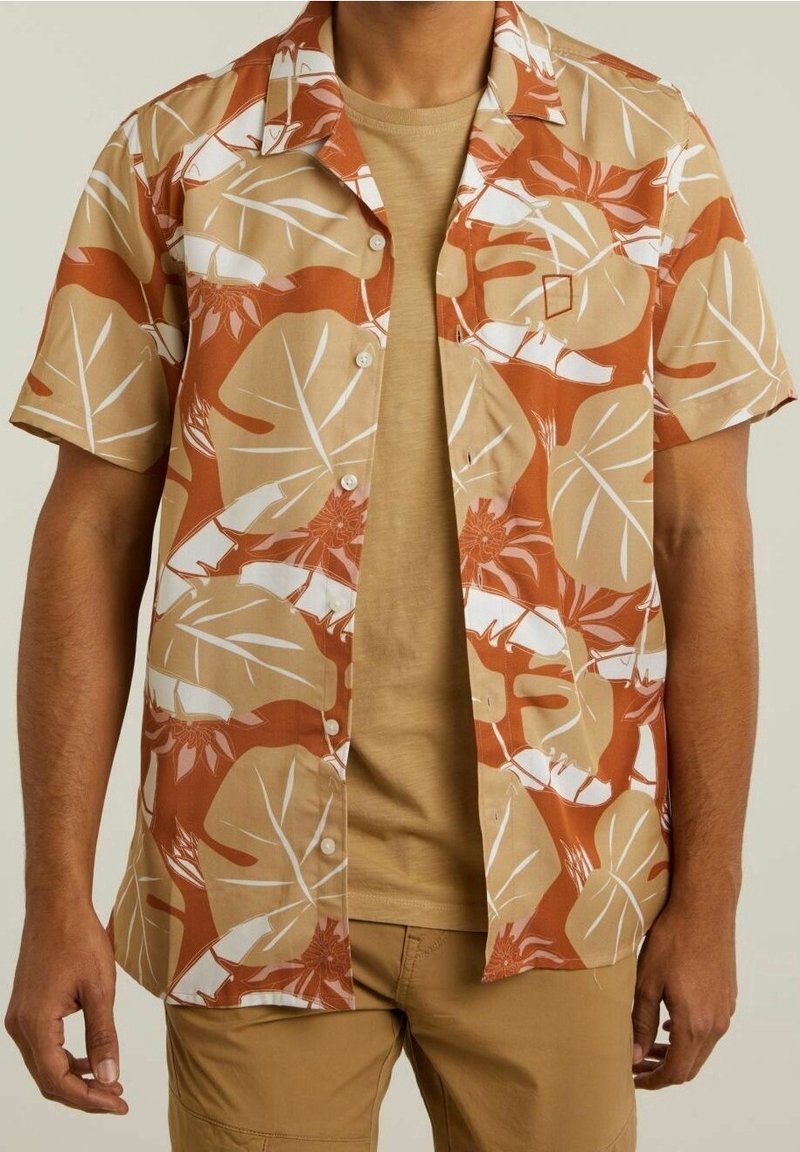 Camicetta a maniche corte beige con un motivo di foglie tropicali marroni e bianche. Presenta una chiusura con bottoni e una vestibilità morbida sopra una maglietta color sabbia.