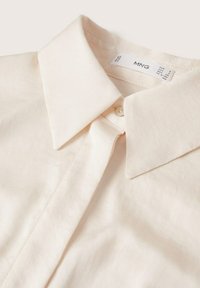 Blouse beige clair avec un col classique, une fermeture à boutons, une texture de tissu douce et une étiquette indiquant la taille et la marque.