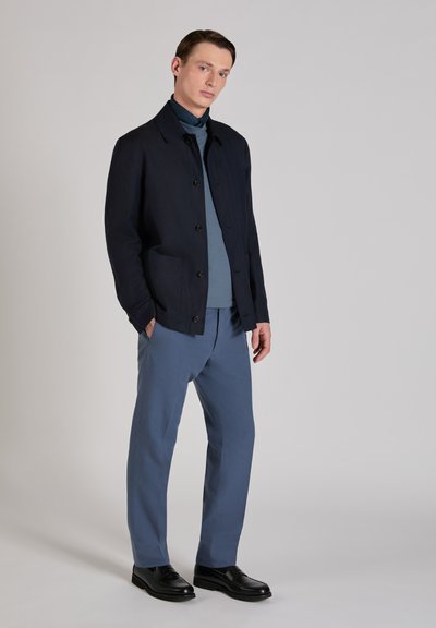 Marineblaue Jacke mit Knöpfen, blauer Rollkragenpullover und hellblaue Hose. Schwarze Schuhe. Glatter Stoff, minimalistisches Design.