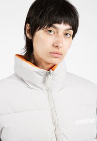 Chaqueta de plumas gris claro con cuello alto y forro naranja, que cuenta con un cierre frontal de cremallera y un logo blanco en el pecho.