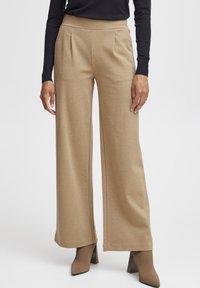 Pantalons larges beige avec une texture lisse, un devant plissé, une taille haute et des lignes épurées, associés à des chaussures à talons dans une teinte complémentaire.
