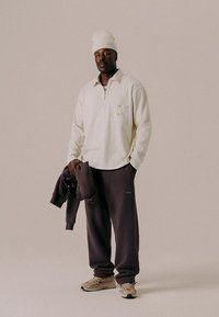 Hvit genser med glidelås og liten brystlomme, mørkebrune joggebukser og beige joggesko; modellen holder en brun hettegenser.