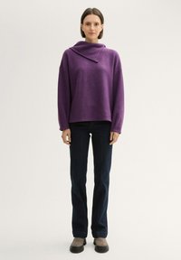 Lila Pullover mit Kapuzenkragen, lockerer Passform und langen Ärmeln. Kombiniert mit dunkelblauen Jeans und grauen Knöchelstiefeln mit schwarzen Sohlen.