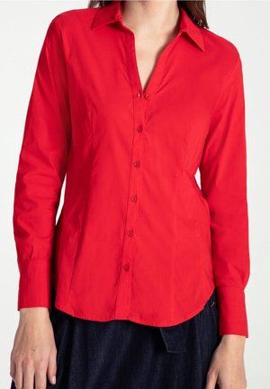 Vrouw draagt een felrode blouse met lange mouwen en kraag met knopen en een donkerblauwe broek tegen een witte achtergrond.