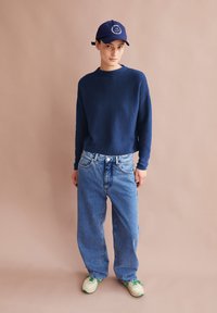 Marineblauer Pullover, hellblaue lässige Jeans, weiße Sneakers mit grünen Akzenten und eine marineblaue Kappe mit einem runden Logo auf der Vorderseite.
