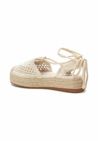Espadrilles de color beige tejido con suela de cuerda gruesa, lados abiertos y lazos ajustables. Parte superior texturizada con un detalle tipo red.