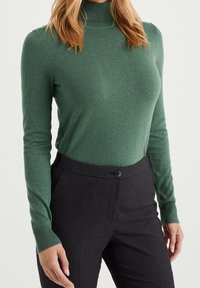 Mörkt grön polo-tröja i mjuk textur, i kombination med mönstrade svarta byxor med enkel knappstängning.