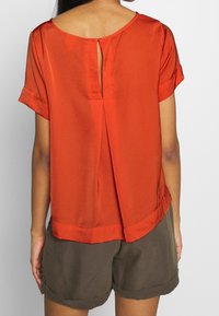 Blusa rossa a maniche corte in tessuto leggero con scollo rotondo, dettaglio a oblò sulla schiena e orlo dritto, abbinata a pantaloni corti color oliva.