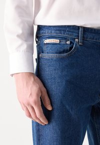 Jeans de denim azul con corte recto, que presentan costuras contrastantes, una pequeña etiqueta de Calvin Klein en rojo y blanco, y detalles metálicos en plata.