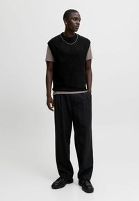Zwarte mouwloze gebreide top over een lichtgrijze T-shirt, gecombineerd met brede zwarte gestreepte broek en zwarte veterschoenen.