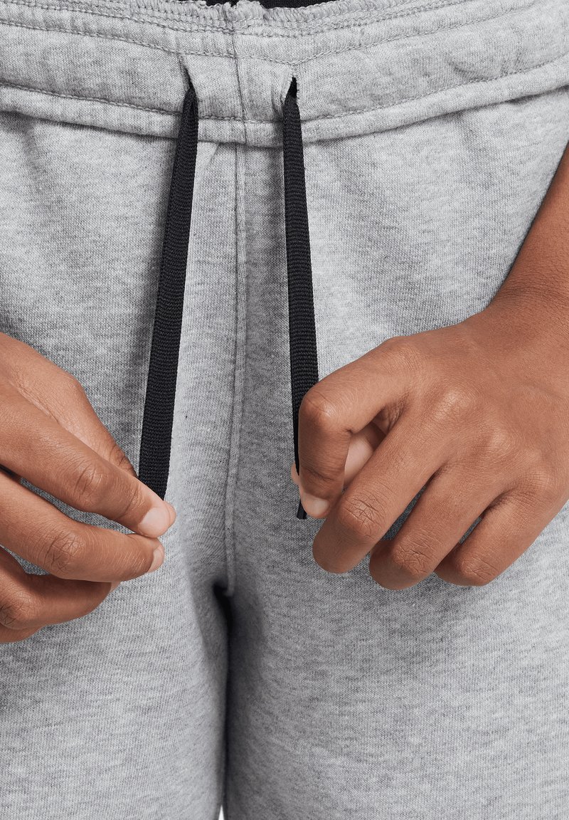 Grijze joggingbroek met een tailleband met trekkoord. De koorden zijn zwart en de stof heeft een zachte, gestructureerde uitstraling. Close-up van handen die de koorden verstellen.
