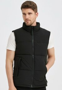 Gilet rembourré noir avec col montant, fermeture éclair à l'avant et poches latérales. Porté par-dessus une chemise blanche, texture lisse, design matelassé.