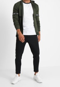 Giacca a zip verde oliva con accenti neri e testo sulle maniche, abbinata a una T-shirt bianca e pantaloni neri a sigaretta. Sneakers bianche.