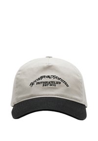 Cappello di stoffa beige con visiera nera; presenta una forma curva e testo ricamato "GOOD FOR NOTHING BRITISH ATELIER EST 2011" sul davanti.