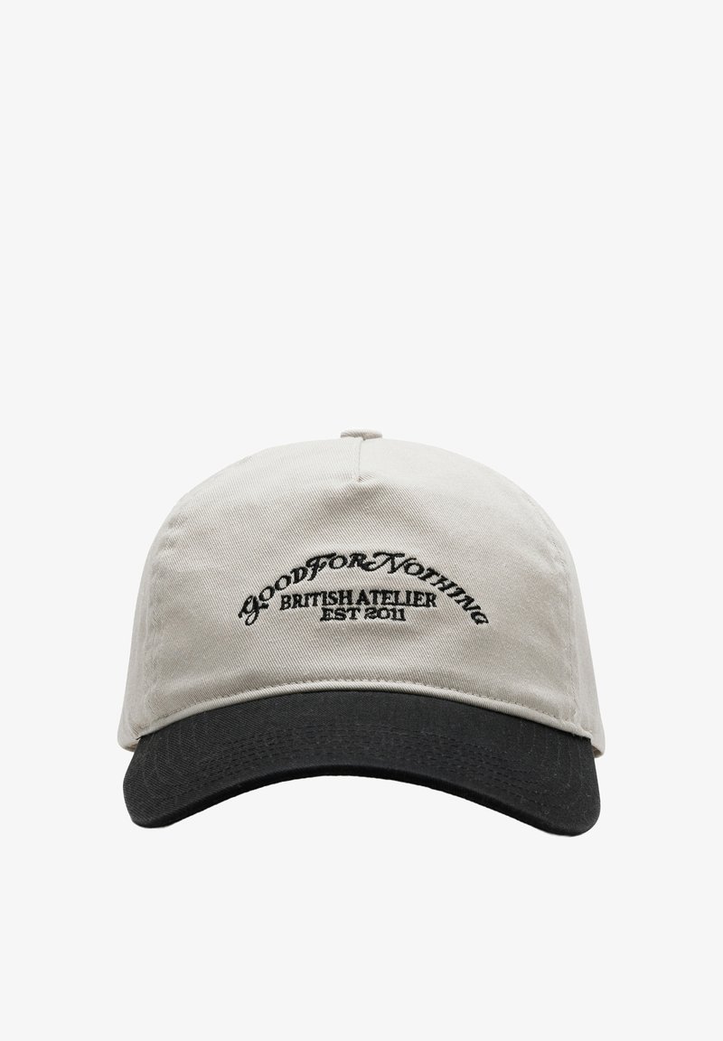 Beige stoffen pet met een zwarte klep; heeft een gebogen vorm en geborduurde tekst "GOOD FOR NOTHING BRITISH ATELIER EST 2011" aan de voorkant.