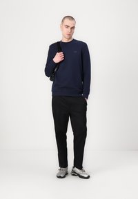 Sudadera azul marino con cuello redondo, puños y dobladillo acanalados, combinada con pantalones negros y zapatillas deportivas plateadas. Los accesorios incluyen un bolso pequeño.