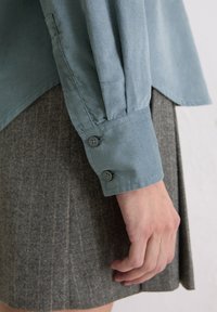 Himmelblaues Longsleeve-Shirt mit knöpfbaren Manschetten, aus weichem Stoff, kombiniert mit einem grauen Nadelstreifenrock. Sichtbare Nähte und Textur.