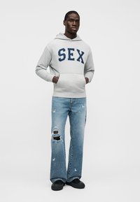 Vyras stovintis kišenėse sukibęs, vilkintis šviesiai pilką džemperį su didelėmis tamsiai mėlynomis raidėmis ir suplyšusiais mėlynais džinsais.