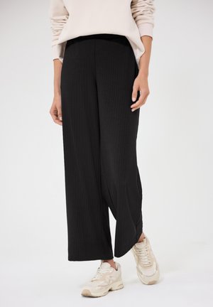 Personne portant un pantalon large en maille côtelée noir, un sweat-shirt beige oversize et des baskets beiges, debout devant un fond clair uni.