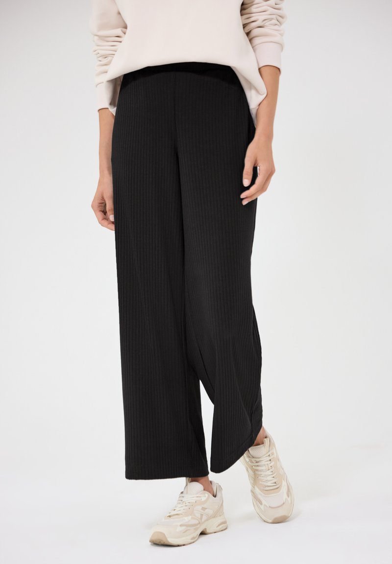 Personne portant un pantalon large en maille côtelée noir, un sweat-shirt beige oversize et des baskets beiges, debout devant un fond clair uni.