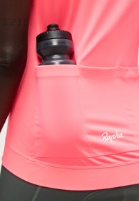 Czarna butelka na wodę w tylnej kieszeni jaskrawo różowego rowerowego trykotu z logo "Rapha", noszona przez osobę o ciemnej karnacji.