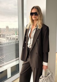 Mujer rubia con traje marrón oscuro grande, camisa clara, gafas de sol, sosteniendo un bolso beige con flecos, de pie junto a una ventana con vista a la ciudad.