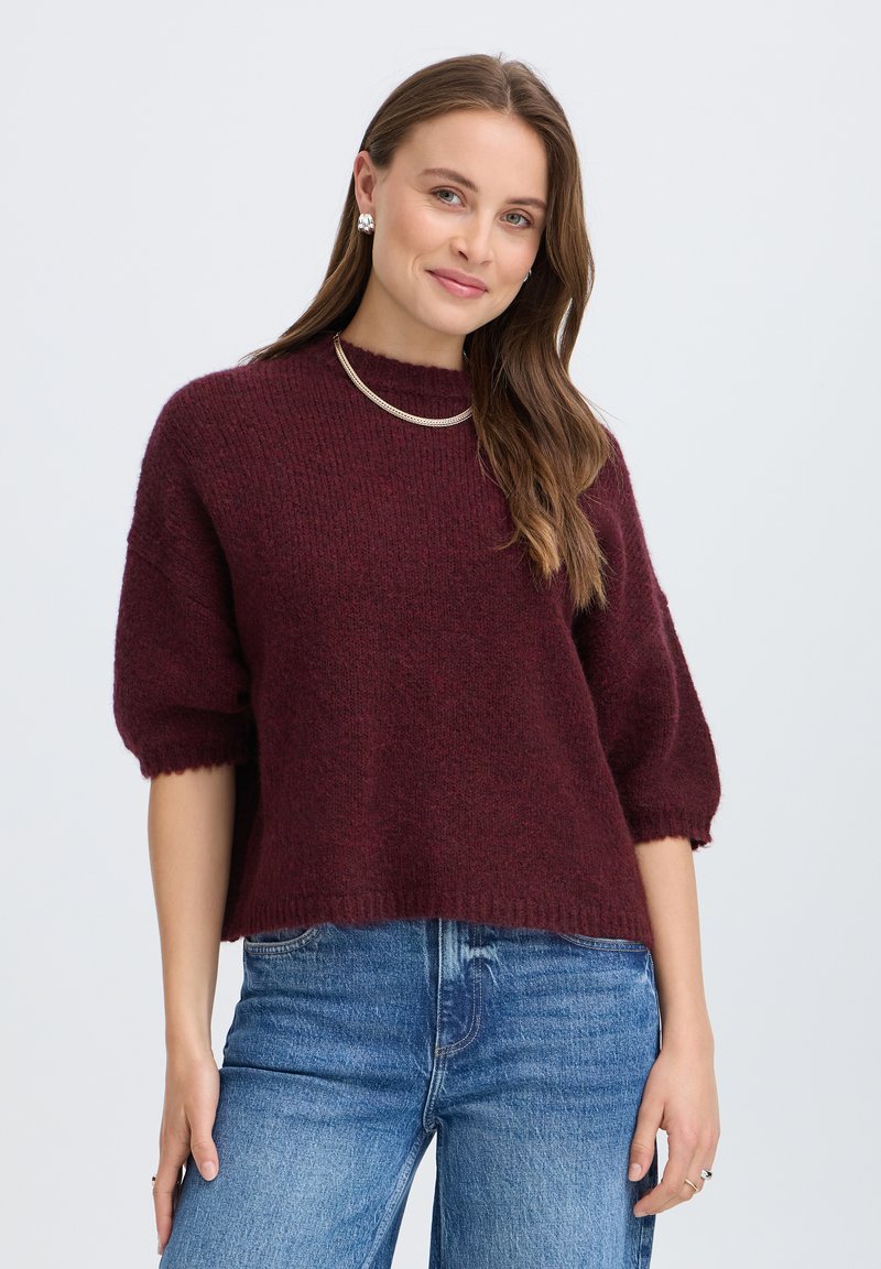 Jeune femme aux longs cheveux bruns portant un pull à manches courtes couleur bordeaux, un jean bleu, un collier en or et des boucles d'oreilles en argent, souriant légèrement.