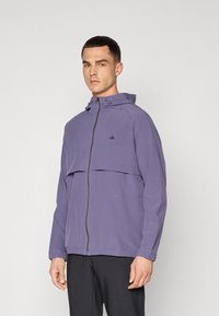 Lilla hettegenser med glidelås foran, med to store brystlommer, elastiske mansjetter og en liten logo på øvre venstre side.
