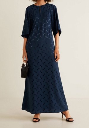 Femme portant une robe bleu marine longueur cheville à pois subtils, tenant un petit sac à main noir et portant des talons ouverts noirs.