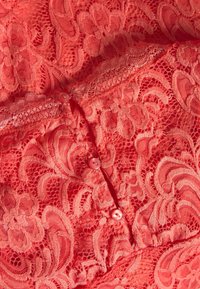 Korálková krajka s květinovými vzory, s vlnitým okrajem a třemi růžovými knoflíky na předním zapínání. Měkčená textura.