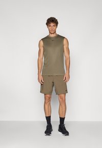 Olivgrünes ärmelloses Sportoberteil, kombiniert mit passenden Shorts; beides aus leichtem Material. Schwarze Sneaker und Socken runden das Outfit ab.