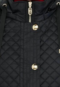 Chaqueta negra acolchada con cuello alto, con cremallera y botones dorados, correas de tela negras y superficie texturizada.