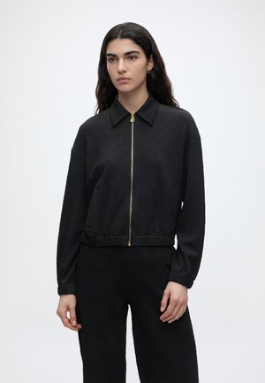 Hosszú sötét hajú nő texturált fekete cipzáras dzsekit gallérral és hozzáillő fekete nadrággal, áll egy sima fehér háttér előtt.