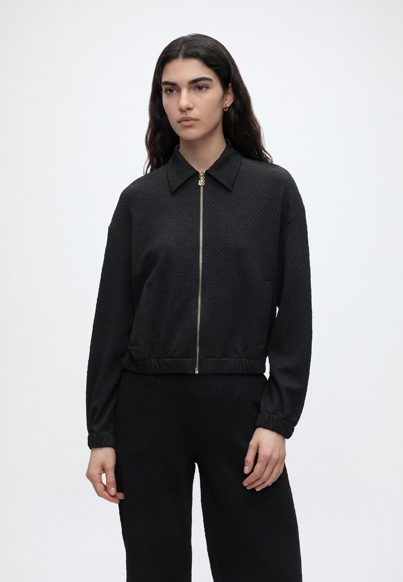 Γυναίκα με μακριά σκούρα μαλλιά φοράει υφασμένη μαύρη ζακέτα με φερμουάρ και γιακά και αντίστοιχο μαύρο παντελόνι, στέκεται μπροστά σε απλό λευκό φόντο.