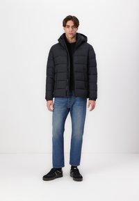 Zwarte pufferjack met capuchon, met stikdetails en een rits aan de voorkant; gedragen over een zwart shirt en gecombineerd met blauwe spijkerbroeken en zwarte sneakers.