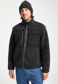 Veste en polaire noire avec un dessus en tissu lisse, poches grises et un col haut. Portée avec un bonnet en tricot noir assorti.