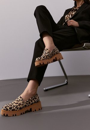 Scarpe slip-on con stampa leopardata, dotate di una suola spessa color tan con bordi frastagliati. Indossate con pantaloni neri, evidenziando un design moderno e una texture interessante.