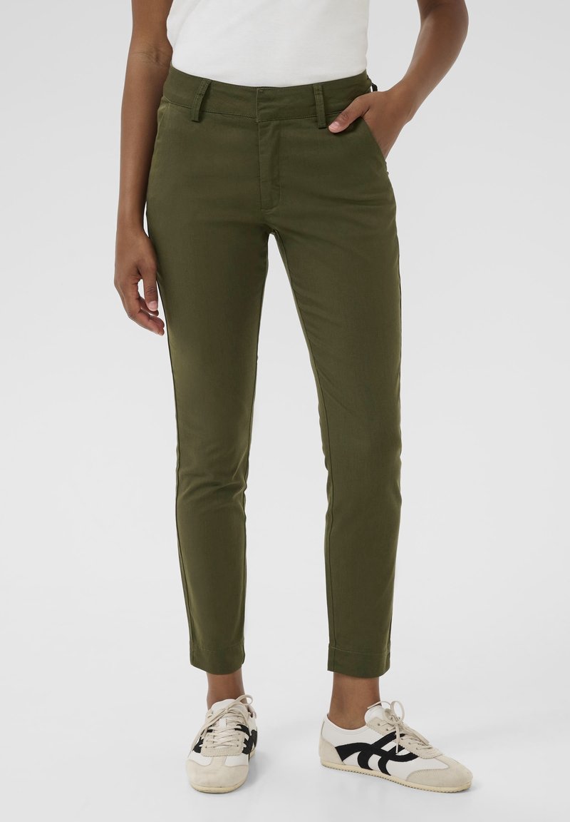 Personne portant un pantalon ajusté vert olive et des baskets blanches avec des détails noirs, debout avec une main dans la poche sur un fond uni.