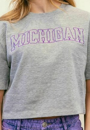 Person iført en grå cropped T-shirt med "MICHIGAN" i lilla blokbogstaver og højtaljede lilla cowboybukser.